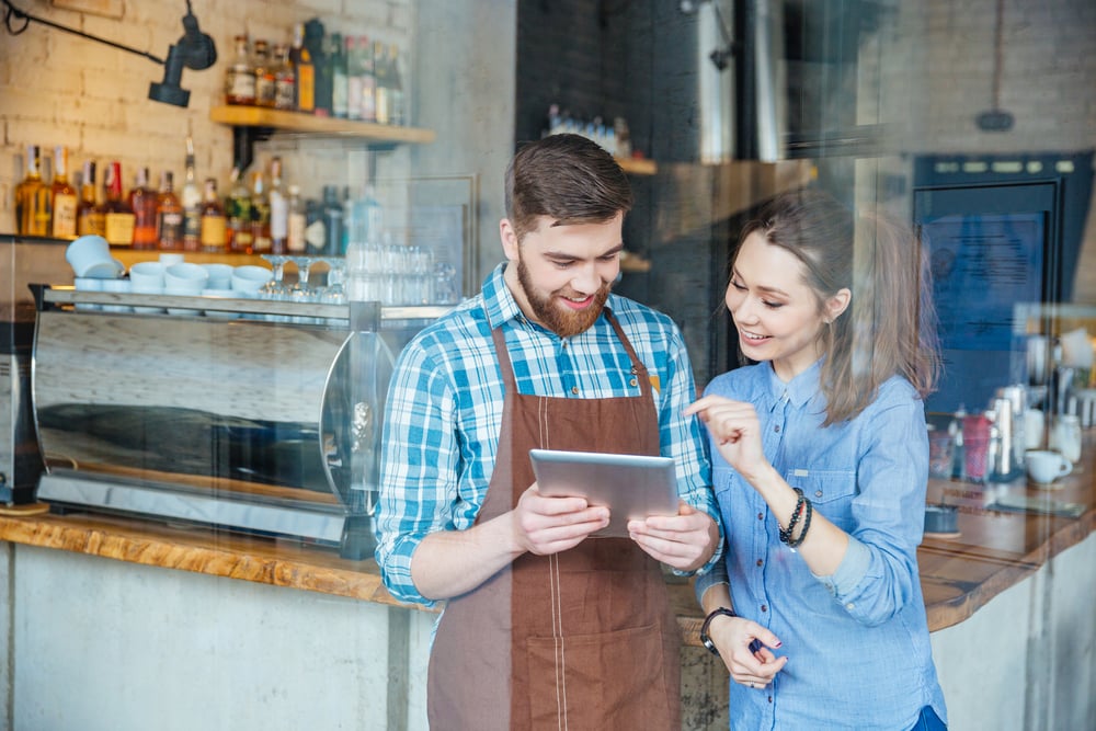 Lächelnd gut aussehend Kellner hält Tablette und junge hübsche Frau zeigt auf sie in Coffee-Shop