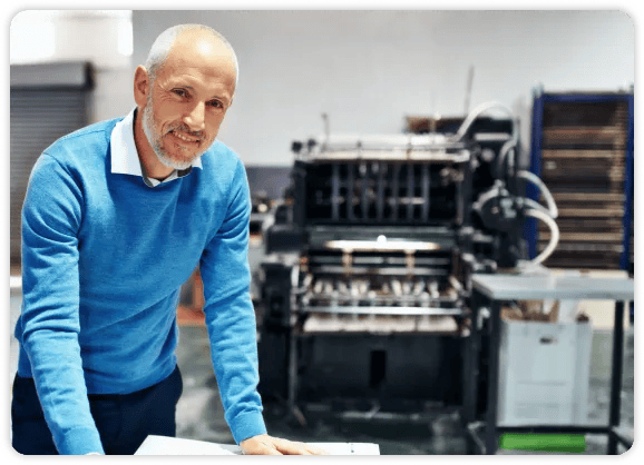 älterer Mann in blauem Hemd vor einer Papierfaltmaschine stehend